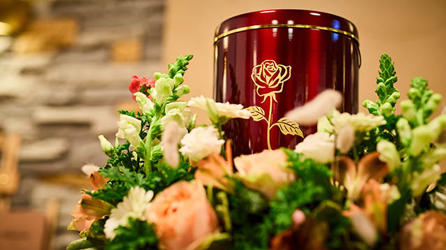 Bestattungsarten: rote Urne mit goldener Rose – Schwarz & Sohn Rote Urne mit goldener Rosenverzierung zwischen Blumenarrangement; Detailaufnahme als Symbol für Urnen- und Feuerbestattung.