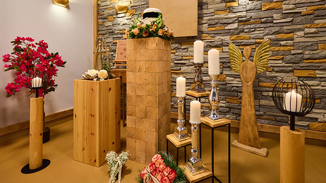 Bestattungsvorsorge – Symbolfoto mit Urnenaufbahrung & Kerzen Urne auf hohem Podest mit Blumen, Kerzenständern und Holzengel vor Steinwand – stimmungsvolle Aufnahme zur Bestattungsvorsorge.
