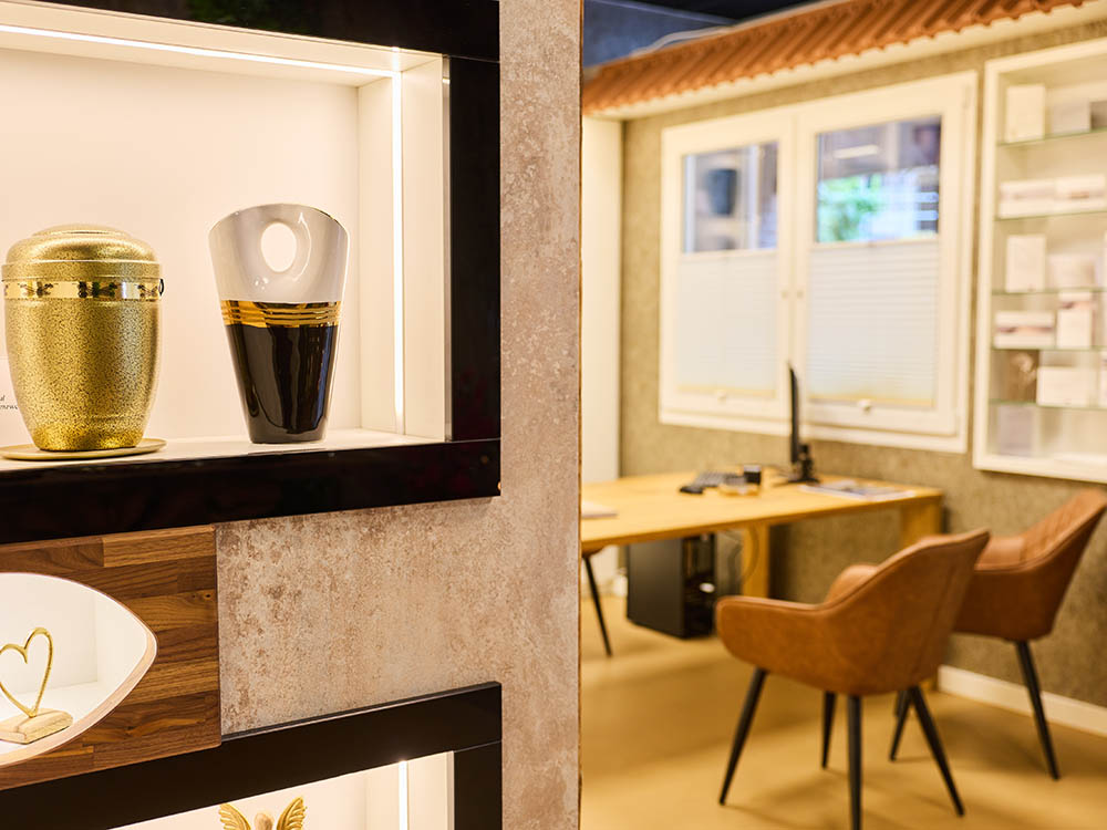 Persönliche Beratung & Organisation – Beratungsraum mit Urnennischen Beleuchtete Nischen mit Urnen im Vordergrund; dahinter Beratungsraum mit Holztisch und zwei Stühlen im Bestattungsinstitut in Versmold.
