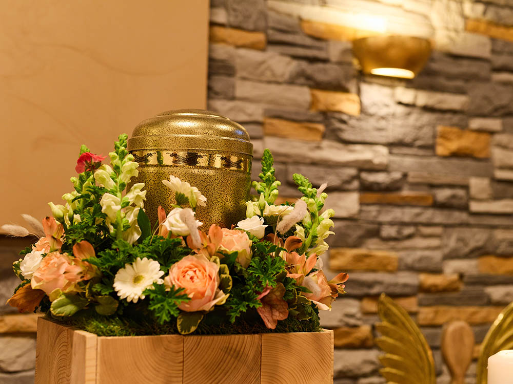 Bestattungsarten: goldene Urne im Blumenarrangement – Schwarz & Sohn Goldene Urne auf Holzpodest mit zartem Blumenarrangement vor Steinwand und warmem Licht – Symbolfoto für Bestattungsarten in Versmold.