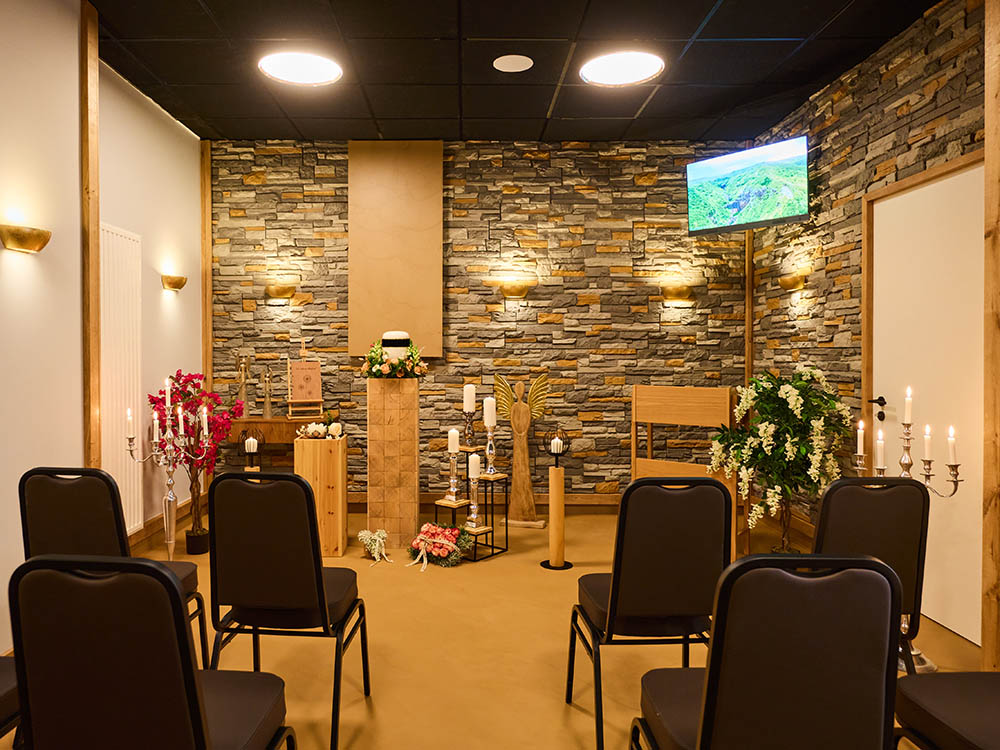 Trauerhalle Versmold – Urnenaufbahrung, Kerzen & Sitzreihen Innenansicht der Trauerhalle von Schwarz & Sohn in Versmold: Blick auf Urnenpodest mit Kerzen vor Steinwand, seitlich Bestuhlung und Bildschirm.